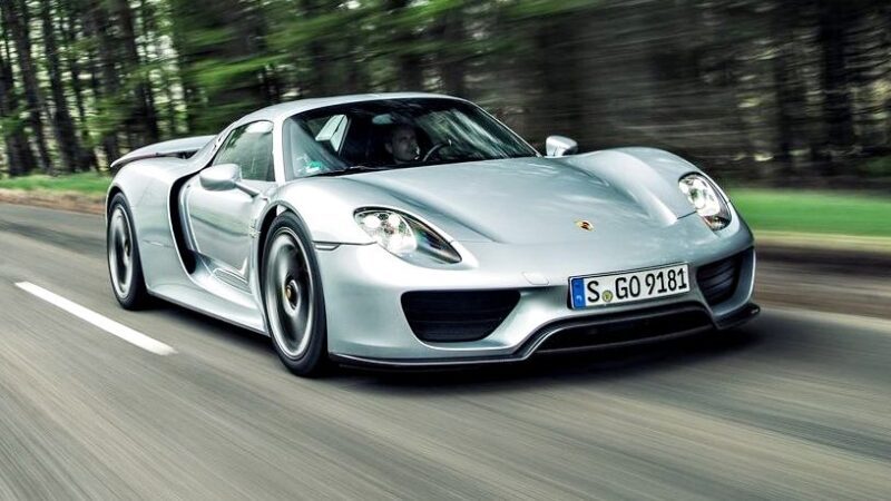 A silver Porsche 918 Spyder sports car speeds down a tree-lined road, its blurred background hinting at dynamic motion—a thrilling scene perfect for fans of the Comparison: Porsche Carrera GT Vs Porsche 918 Spyder debate.