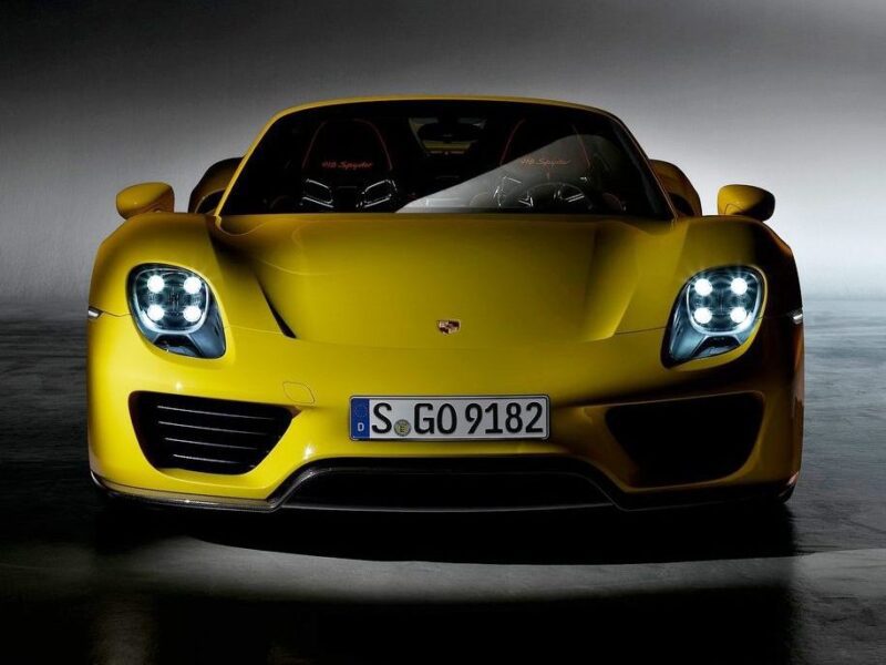 A yellow Porsche sports car viewed from the front with headlights on and a German license plate reading "S GO 9182"—a striking image for any Comparison: Porsche Carrera GT Vs Porsche 918 Spyder enthusiast.
