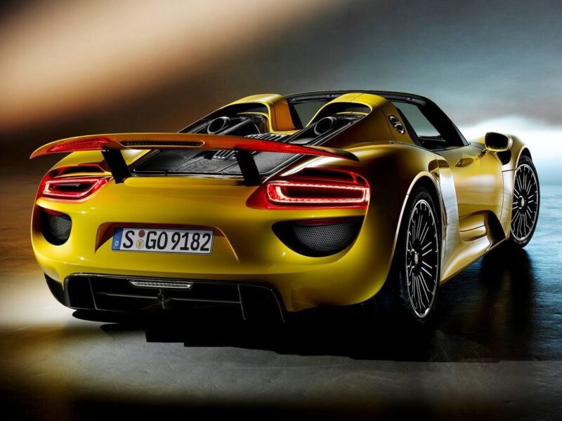 A yellow Porsche 918 Spyder sports car is shown from the rear, its taillights illuminated and large rear wing raised, highlighting its advanced aerodynamics in a Comparison: Porsche Carrera GT Vs Porsche 918 Spyder.