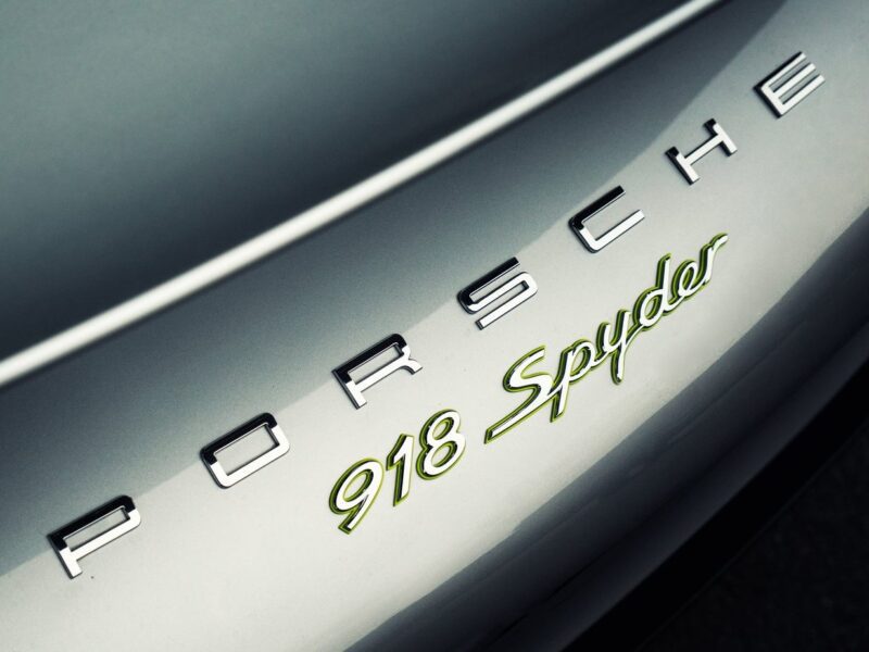 Close-up of a Porsche 918 Spyder logo on the rear of a silver car, highlighting "Porsche" in chrome and "918 Spyder" in green and gold—a striking detail for any Comparison: Porsche Carrera GT Vs Porsche 918 Spyder.