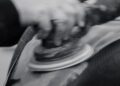 A person uses an electric sander to smooth the surface of a carbon fiber material, preparing it for use in an exotic car like the Porsche 963 RSP.