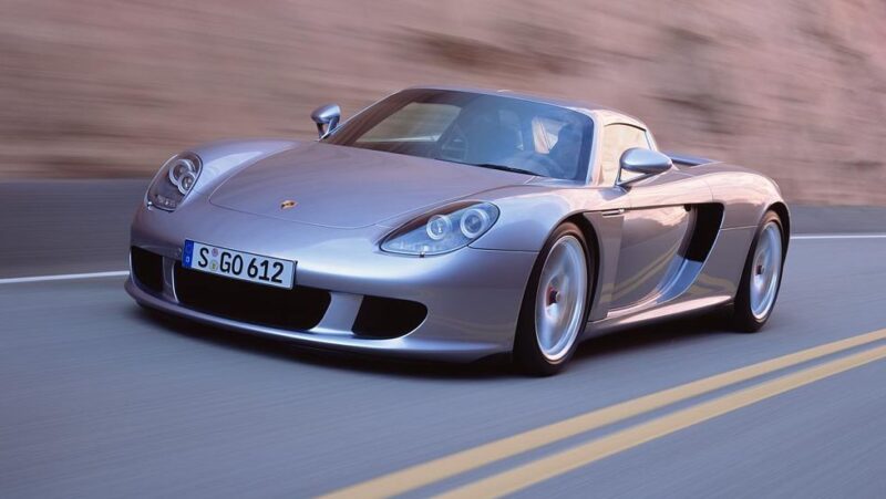 A silver Porsche Carrera GT sports car drives on a winding road with a blurred rocky background, showcasing its iconic design—a timeless contrast in the ongoing debate: Porsche Carrera GT vs Porsche 918 Spyder.