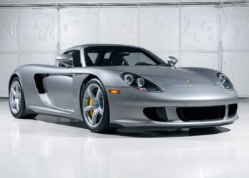 A silver Porsche Carrera GT is parked indoors in front of a white industrial-style wall, showcasing Stuttgart engineering at its finest.