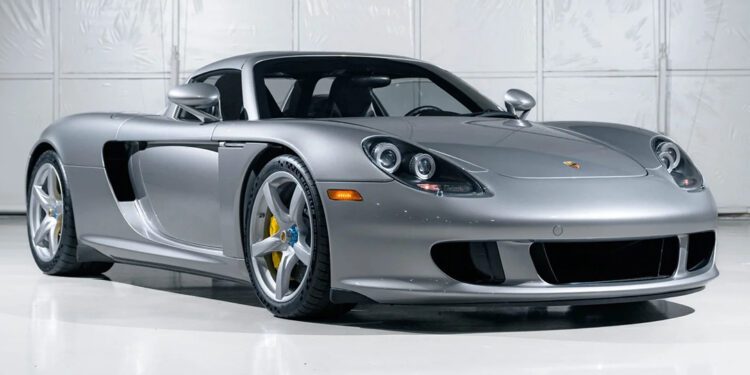 A silver Porsche Carrera GT is parked indoors in front of a white industrial-style wall, showcasing Stuttgart engineering at its finest.