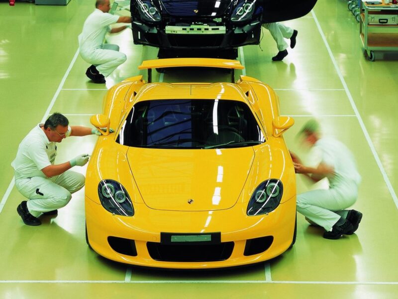 Three workers in white uniforms inspect and clean a yellow sports car on a glossy, organized factory floor, reminiscent of the precision seen in the Comparison: Porsche Carrera GT Vs Porsche 918 Spyder.