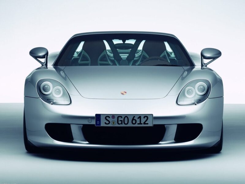 Front view of a silver Porsche sports car with headlights on and a visible license plate reading "S GO 612" against a plain background, perfect for a Comparison: Porsche Carrera GT Vs Porsche 918 Spyder showcase.