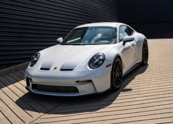 A white Porsche sports car from Porsche Centre Langley is parked on a paved, striped surface next to a dark metal wall under daylight—an eye-catching dealer spotlight for exotics for sale.