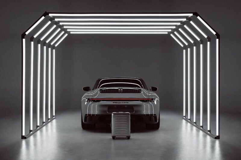 A silver sports car is parked under a modern, illuminated frame structure with a Porsche Design Roadster Aluminum Luggage suitcase placed behind the vehicle, inviting you to buy into ultimate sophistication.