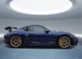 A Gentian Blue Porsche 718 Cayman GT4 RS with the Weissach Package and gold rims, side view, parked in a modern, circular indoor space with a large skylight overhead.