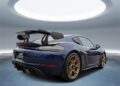 A Porsche 718 Cayman GT4 RS in striking Gentian Blue with the Weissach Package, gold wheels, and a large rear spoiler is parked indoors beneath a circular skylight.