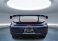 Rear view of a Gentian Blue Porsche 718 Cayman GT4 RS with the Weissach Package, featuring a large rear wing, displayed indoors under a circular skylight.