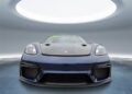 A front view of a Gentian Blue Porsche 718 Cayman GT4 RS with the Weissach Package is displayed in a modern, circular room with a large skylight above.