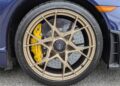 Close-up of a Porsche 718 Cayman GT4 RS wheel with bronze alloy rim, yellow brake caliper labeled "Porsche," and ventilated disc brake, featuring the Weissach Package, parked on a paved surface.