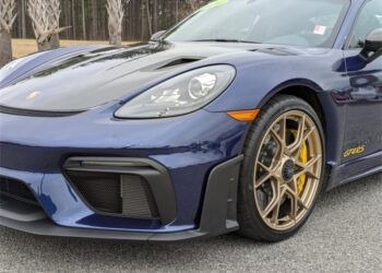 A close-up of the front left side of a Gentian Blue Porsche 718 Cayman GT4 RS sports car, featuring the headlight, wheel, and part of the hood with highlights from the Weissach Package.
