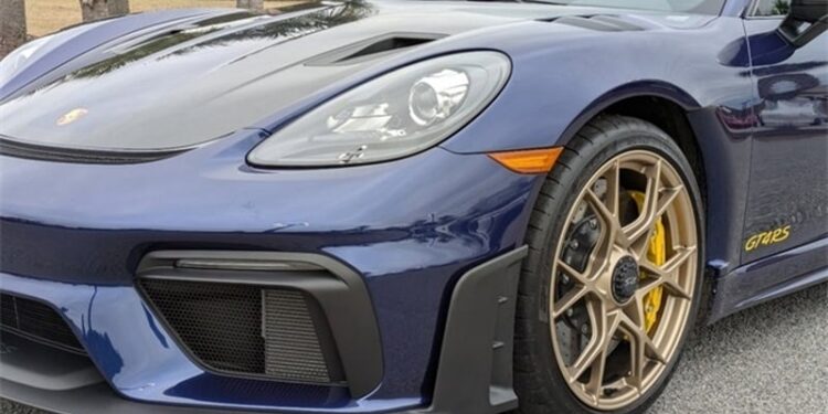 A close-up of the front left side of a Gentian Blue Porsche 718 Cayman GT4 RS sports car, featuring the headlight, wheel, and part of the hood with highlights from the Weissach Package.