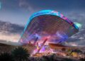 A futuristic, illuminated music festival stage with a large curved roof soars like this upcoming F1 track’s opening corner—20 storeys up in the sky—aglow with colorful lights, fireworks, and a vibrant crowd of people at night.