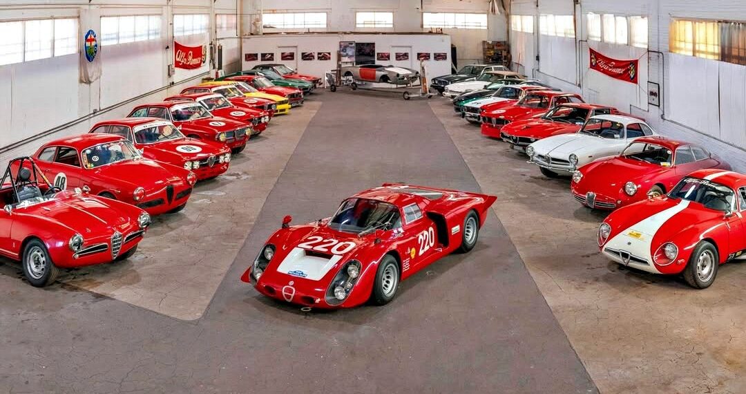 A collection of classic sports cars, mostly red, arranged in a large garage with a race car featuring number 220 in the center, showcased as part of the RM Sotheby’s Quadrifoglio Collection Monterey 2025.