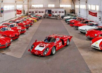 A collection of classic sports cars, mostly red, arranged in a large garage with a race car featuring number 220 in the center, showcased as part of the RM Sotheby’s Quadrifoglio Collection Monterey 2025.