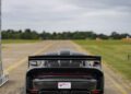 A black supercar is parked on a deserted runway under a “Supercar Fest” banner, with green fields and cloudy skies in the background.