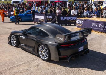 A black sports car with a large rear wing is displayed at an outdoor car event, surrounded by spectators and other parked cars.