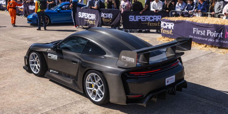 A black sports car with a large rear wing is displayed at an outdoor car event, surrounded by spectators and other parked cars.