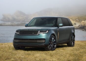 A green Range Rover SV Carmel Edition is parked on grass near a rocky shoreline, with fog and hills in the background—a true highlight among Exclusive Rovers.