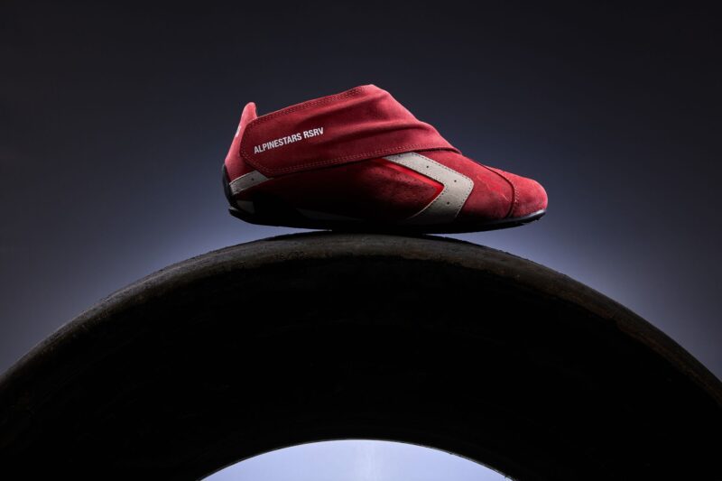 A red Alpinestars RSRV racing shoe with white accents rests on top of a black tire against a dark gradient background, showcasing its F1 DNA.