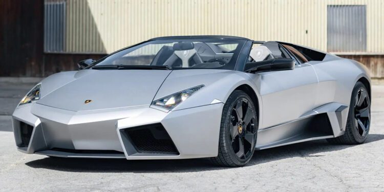 A sleek Lamborghini Revénton Roadster sports car with an angular design is parked on a concrete surface in front of a corrugated metal building, reminiscent of an exclusive find at an RM Sothebys Milan Auction.