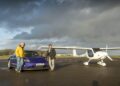 An Emotional Homecoming: James May and Richard Hammond stand on a wet runway—one leaning on a blue electric car, the other by a small white propeller plane—as they race to the Top Gear track in EVs under a cloudy sky.
