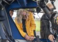 Three people examine the controls inside a helicopter cockpit, with one person in a yellow jacket leaning in closely—capturing the anticipation of an emotional homecoming like James May and Richard Hammond racing to the Top Gear track in EVs.