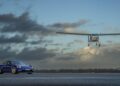 A blue sports car is parked on a runway as a small white airplane descends to land, evoking an emotional homecoming as James May and Richard Hammond race to the Top Gear Track in EVs beneath cloudy skies.