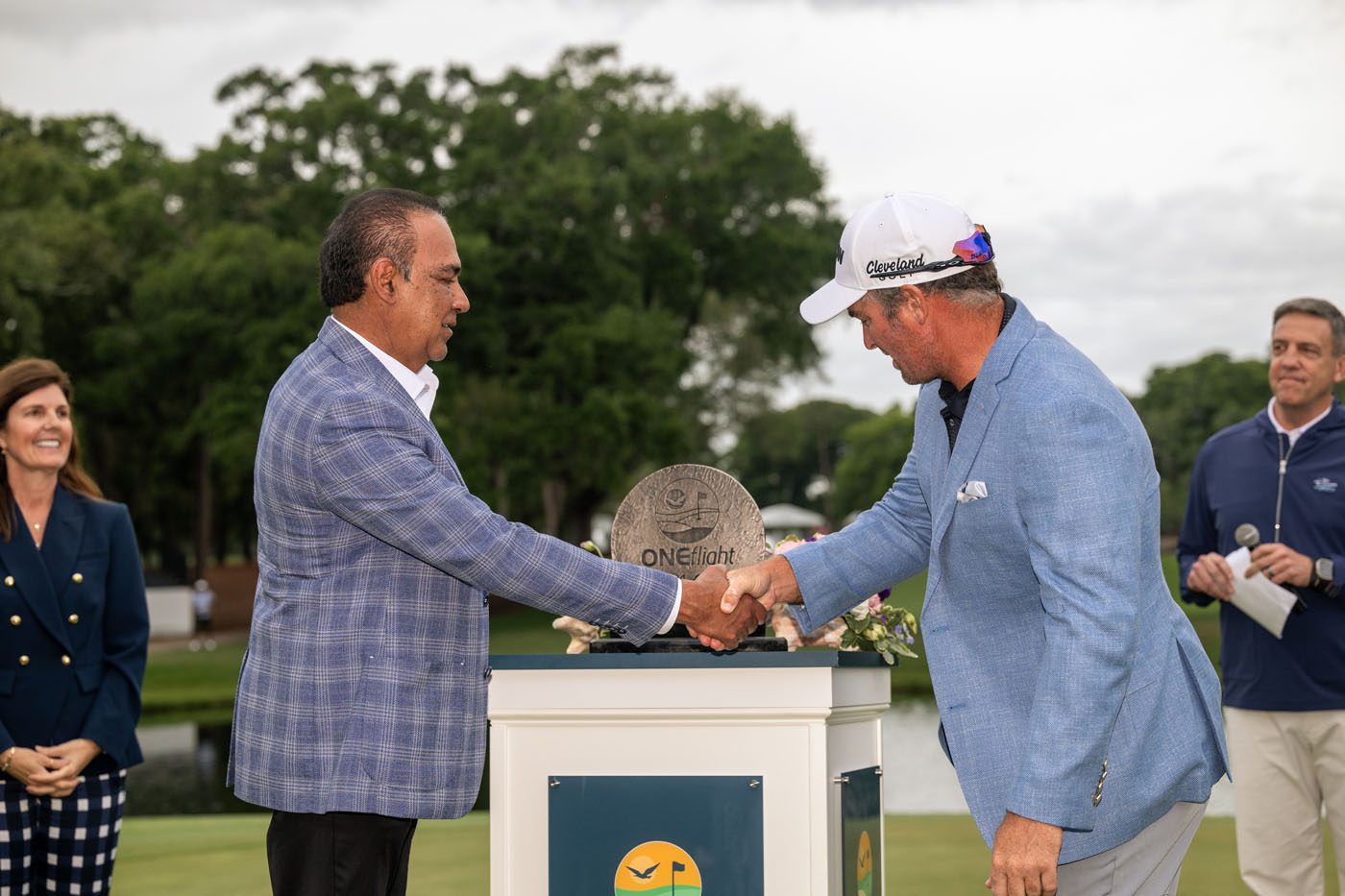 Two men in blazers shake hands over a trophy on a podium outdoors, with several people watching in the background.