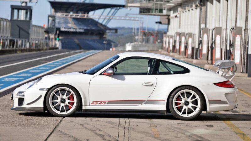 A white Porsche 911 GT3 RS 4.0, the last production Porsche with a naturally-aspirated Mezger flat-six engine, is parked in a pit lane at a racetrack, with garages and grandstands visible in the background.