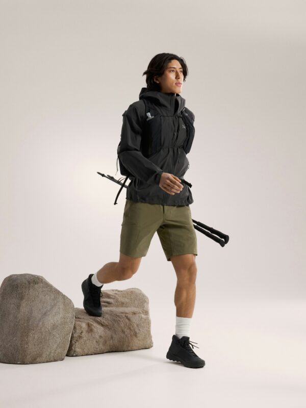A person wearing a black jacket, green shorts, and black shoes steps over rocks with trekking poles—showcasing essential gear for any outdoor adventure—set against a plain light background.