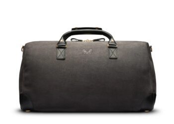 A black leather duffel bag with two handles, a small butterfly logo on the front, and reinforced leather corners—upgrade your style with this elegant piece. The perfect choice among luxury accessories, shown against a white background.