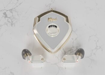 A pair of white and gold wireless earbuds with gray ear tips are placed in front of a matching shield-shaped charging case on a white marble surface, offering a true style upgrade for those seeking luxury accessories.