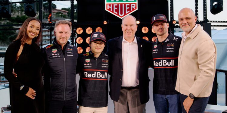 Six people pose together in front of a TAG Heuer sign, with two wearing Red Bull racing gear, at an outdoor Monaco Grand Prix event surrounded by lights and equipment.