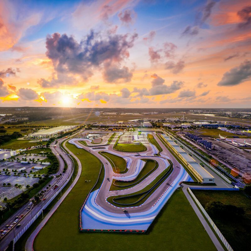 Aerial view of a modern race track complex at sunset, with winding asphalt circuits, green infields, parking areas, and surrounding buildings.