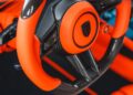 Close-up of a luxury car's orange and black carbon fiber steering wheel with control buttons and paddle shifters, highlighting TECHART performance upgrades for the 992.2 Carrera.