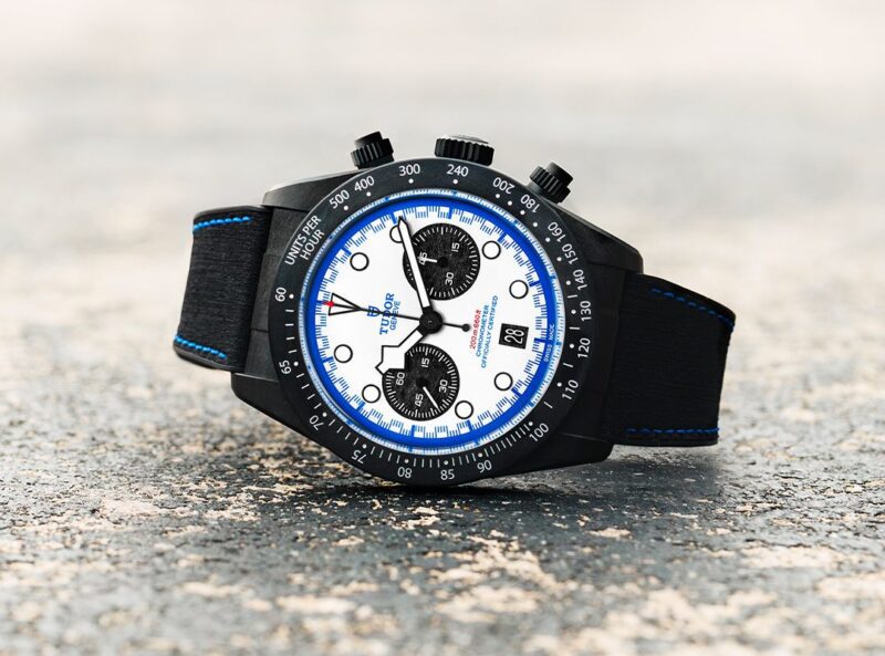 A TUDOR Black Bay Chrono wristwatch with a black case, white dial, black subdials, blue accents, and a black leather strap lies on a textured surface, capturing the dynamic spirit of the Miami F1.