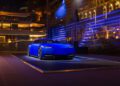 A sleek, futuristic blue car is displayed on a platform under spotlights at night, with modern buildings illuminated in the background.