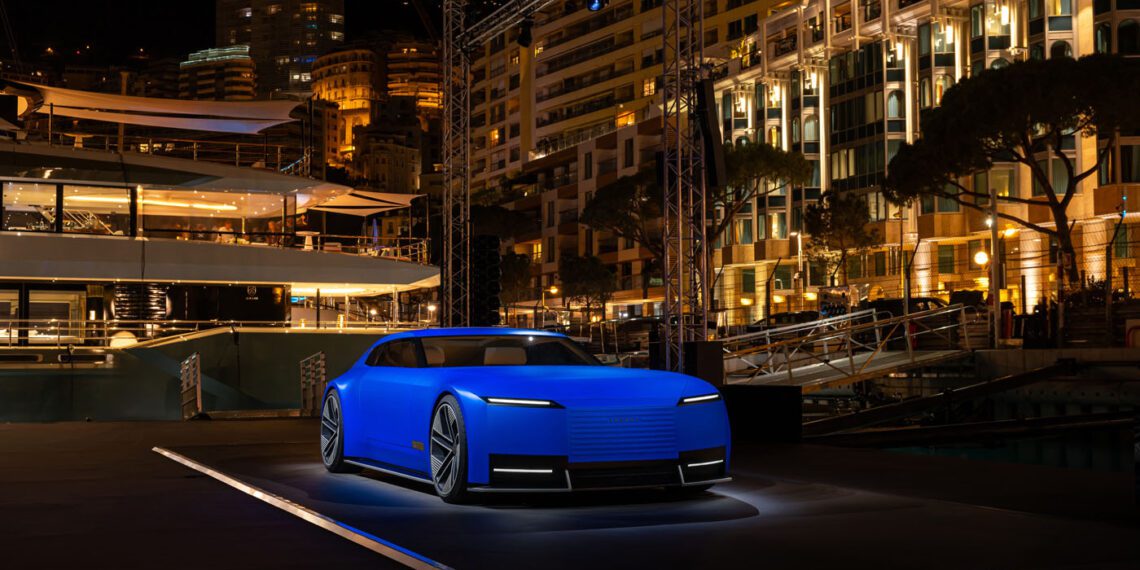 A modern, matte blue concept car is displayed on a lit platform at an outdoor nighttime event with illuminated buildings in the background.