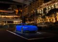 A modern, matte blue concept car is displayed on a lit platform at an outdoor nighttime event with illuminated buildings in the background.