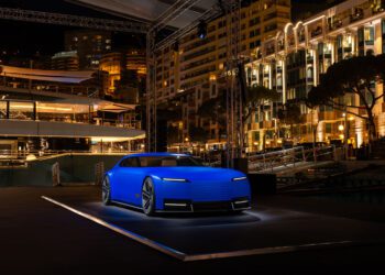 A modern, matte blue concept car is displayed on a lit platform at an outdoor nighttime event with illuminated buildings in the background.