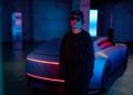 A person wearing dark clothing, sunglasses, and a hat stands in front of a futuristic Jaguar with red and blue neon lights in an industrial Tokyo Art Installation.