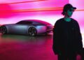 A person in dark clothing and sunglasses stands in front of a silver futuristic car with a sleek design, set against a brightly lit pink and orange background.