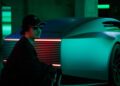 A person wearing sunglasses and a cap sits beside a futuristic car in a dimly lit room with teal and red lights.