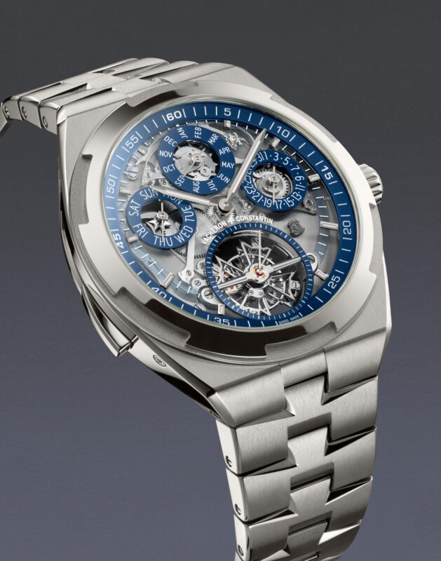 A stainless steel luxury Vacheron Constantin wristwatch with a blue and silver openface skeleton dial, displaying multiple subdials for day, month, and time, on a matching metal bracelet.