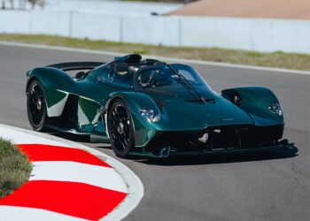 A dark green Aston Martin Valkyrie roadster, with exposed green carbon accents, speeds along a racetrack, rounding a corner as the red and white curb comes into view in the foreground.