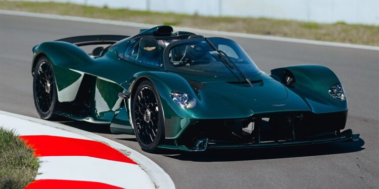 A dark green Aston Martin Valkyrie roadster, with exposed green carbon accents, speeds along a racetrack, rounding a corner as the red and white curb comes into view in the foreground.